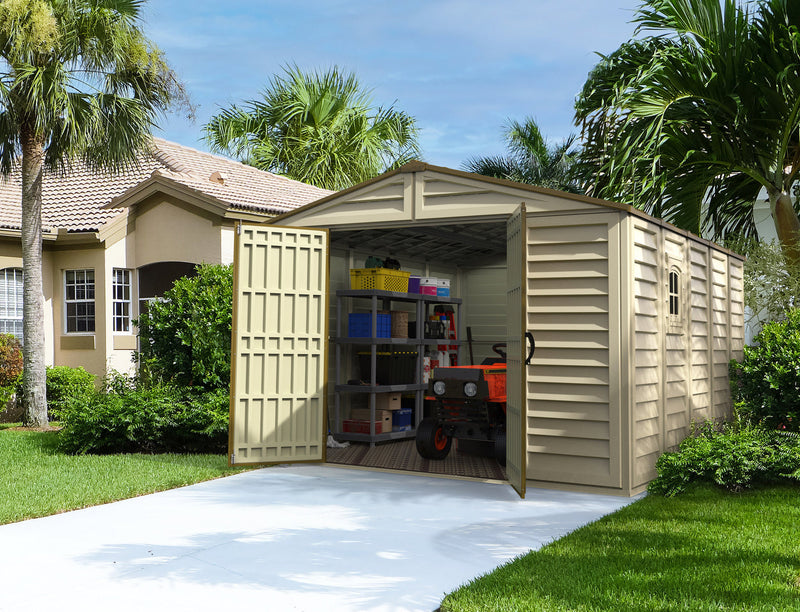 Large beige Duramax Woodbridge Plus 10.5'x13' shed, open and full of tools and storage shelves, set in a lush green yard beside a residential home.