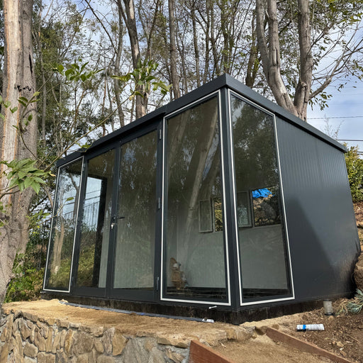 Glass-panel variant of Duramax 13'x10' Insulated Glass Building perched on a raised stone platform in a wooded area.