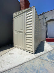 Urban setup of Duramax SideMate Shed with Foundation 4'x8' (06625) placed on a concrete surface beside a gray building and red brick column.