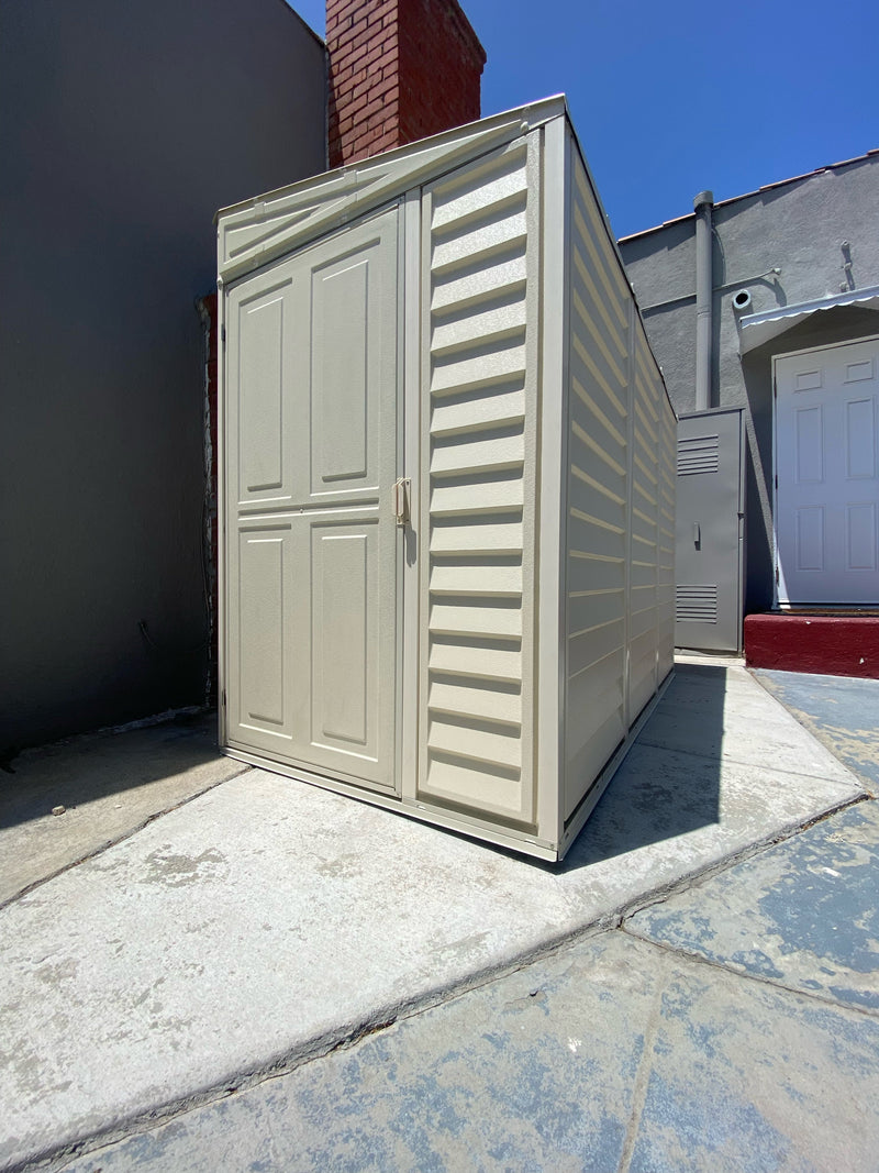 Urban setup of Duramax SideMate Shed with Foundation 4'x8' (06625) placed on a concrete surface beside a gray building and red brick column.